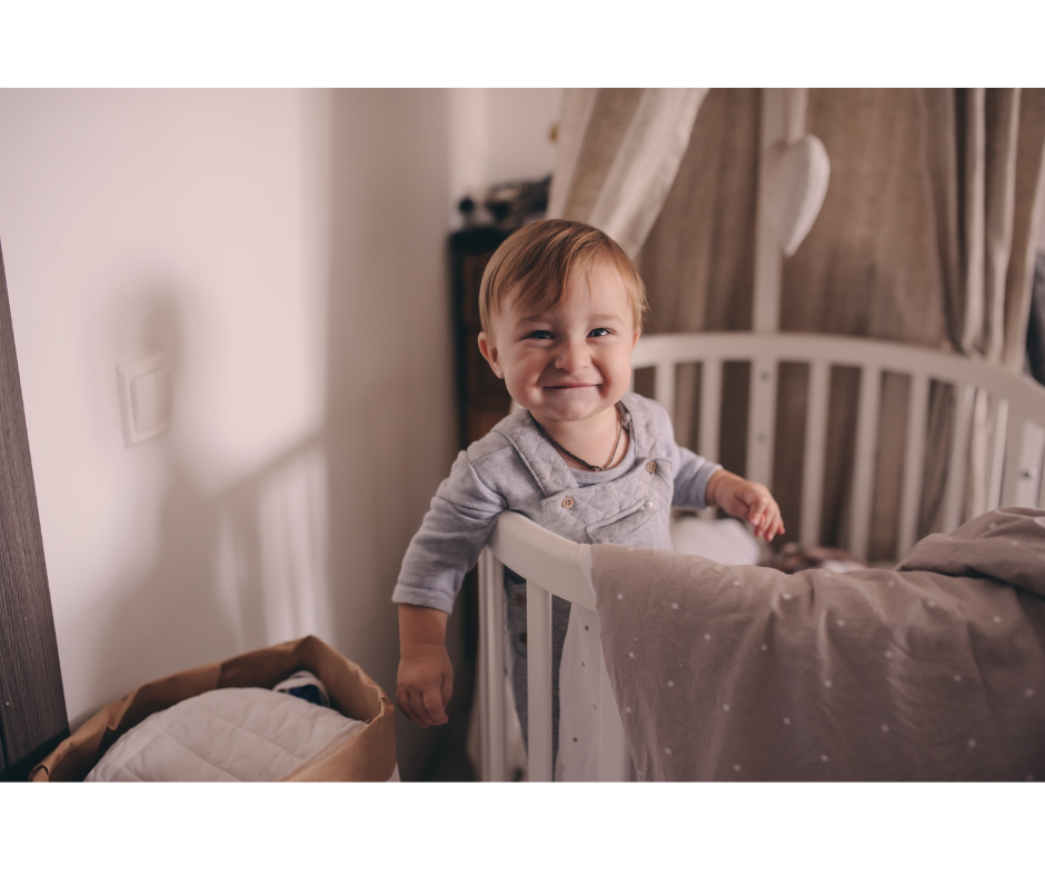 Baby in crib smiling cheekily, in early morning sunlight. Probably awake too early.